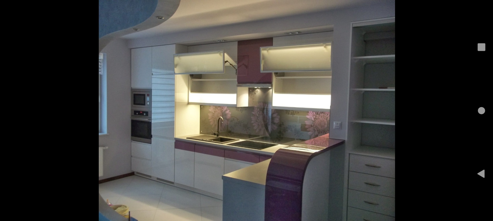 Nowoczesna kuchnia z białymi szafkami, fioletowymi akcentami i podświetleniem LED, widok na zlewozmywak, piekarnik i otwarte półki.