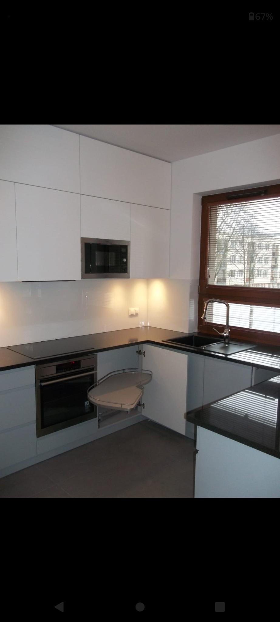 Nowoczesna kuchnia z białymi szafkami, czarnym blatem i narożną półką obrotową, widok na budynek przez okno z żaluzjami.