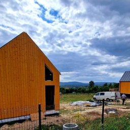 D&W - Dwa drewniane domy w stanie surowym zamkniętym, z widoczną konstrukcją szkieletową i elewacją z desek, na tle pochmurnego nieba i krajobrazu zalesionych wzgórz. Na pierwszym planie ogrodzenie...