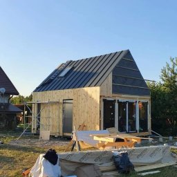 D&W - Wznoszenie domu z drewnianą elewacją, widoczne rusztowanie i częściowo ułożone pokrycie dachu z oknem dachowym.