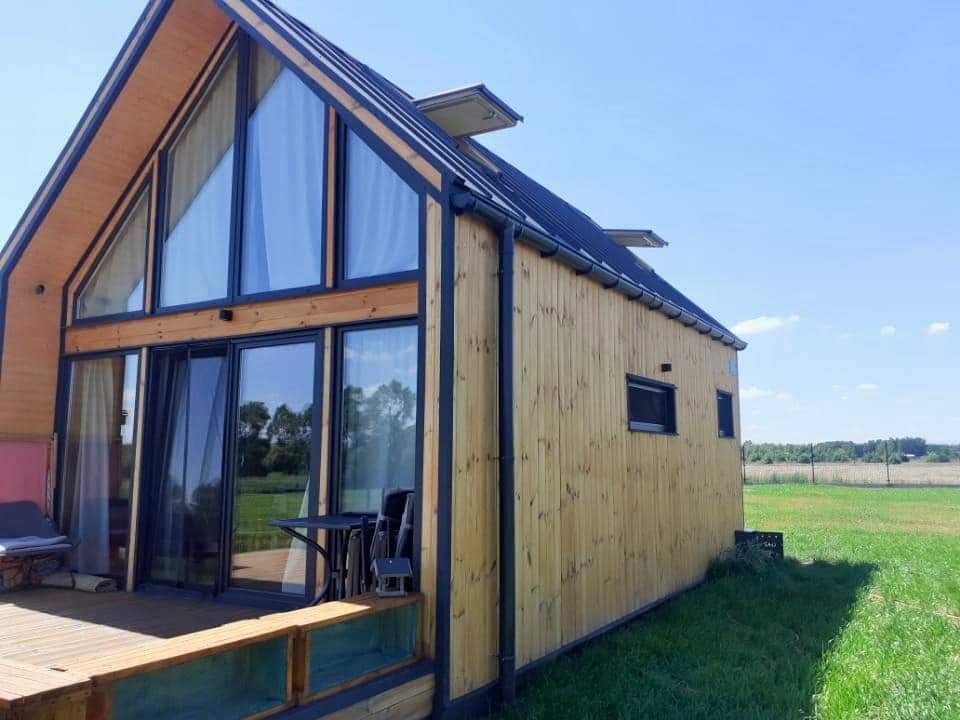 Nowoczesny, drewniany dom z dużymi oknami i tarasem, usytuowany na zielonej łące w słoneczny dzień.