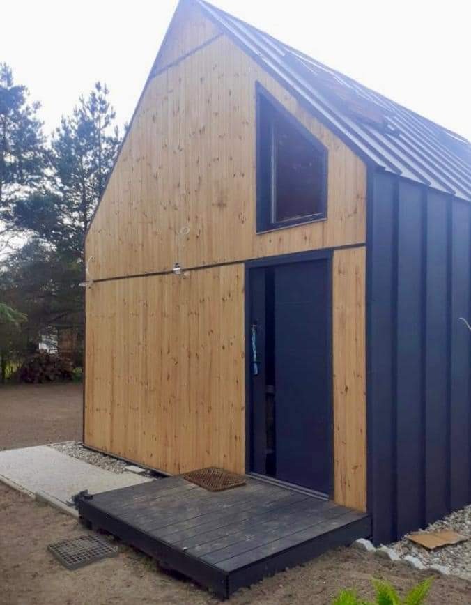 Nowoczesny dom z drewna i blachy na rąbek stojący, z trójkątnym oknem na poddaszu, drewnianym tarasem i chodnikiem z płyt betonowych.