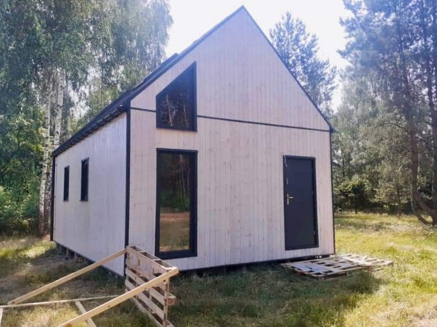 Surowy, drewniany dom w stylu skandynawskim z czarnymi ramami okiennymi i drzwiami, otoczony zielenią, z paletami budowlanymi w pierwszym planie.