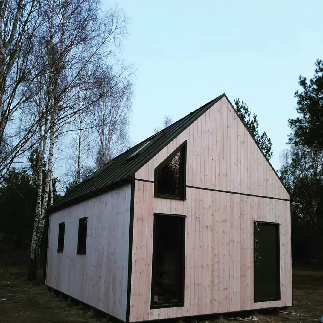 Surowy dom szkieletowy z jasnego drewna, z czarnymi ramami okiennymi i drzwiami, na tle drzew bezlistnych, widziany z boku.