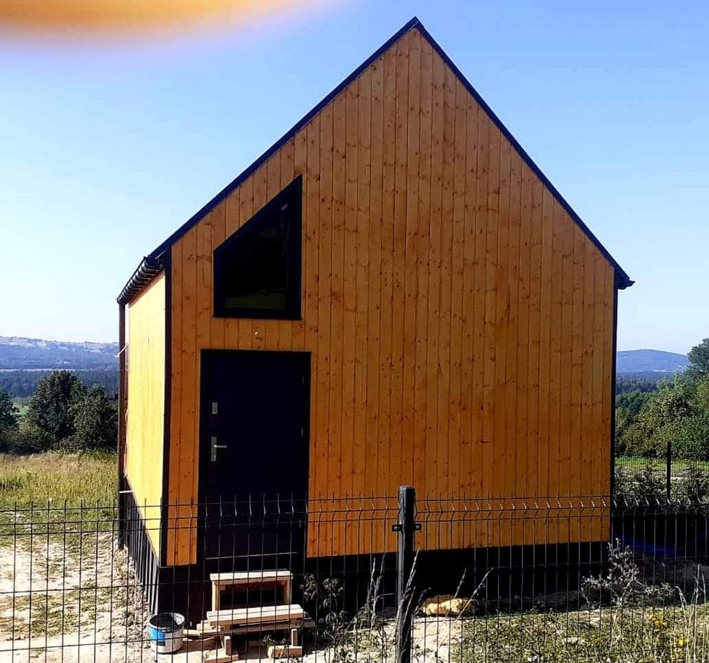 Mały, nowoczesny dom z drewnianą elewacją w naturalnym odcieniu, z czarną stolarką okienną i drzwiową oraz widocznym fragmentem fundamentów, otoczony siatką ogrodzeniową na tle zielonego krajobrazu.