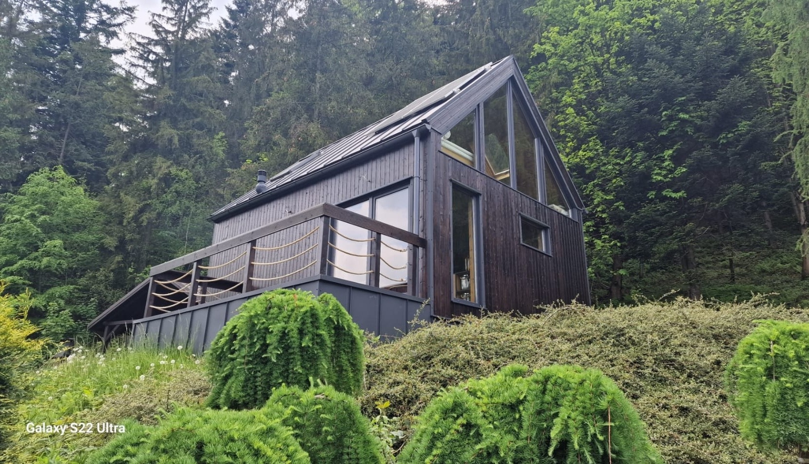 Nowoczesny dom z ciemnego drewna z dużymi oknami i balustradą z lin, wkomponowany w zalesione wzgórze.