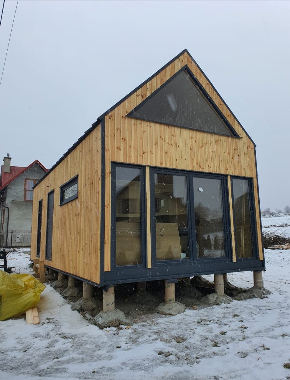 Surowy, drewniany budynek z dużymi oknami i trójkątnym oknem na szczycie, posadowiony na betonowych słupach w zimowym krajobrazie.