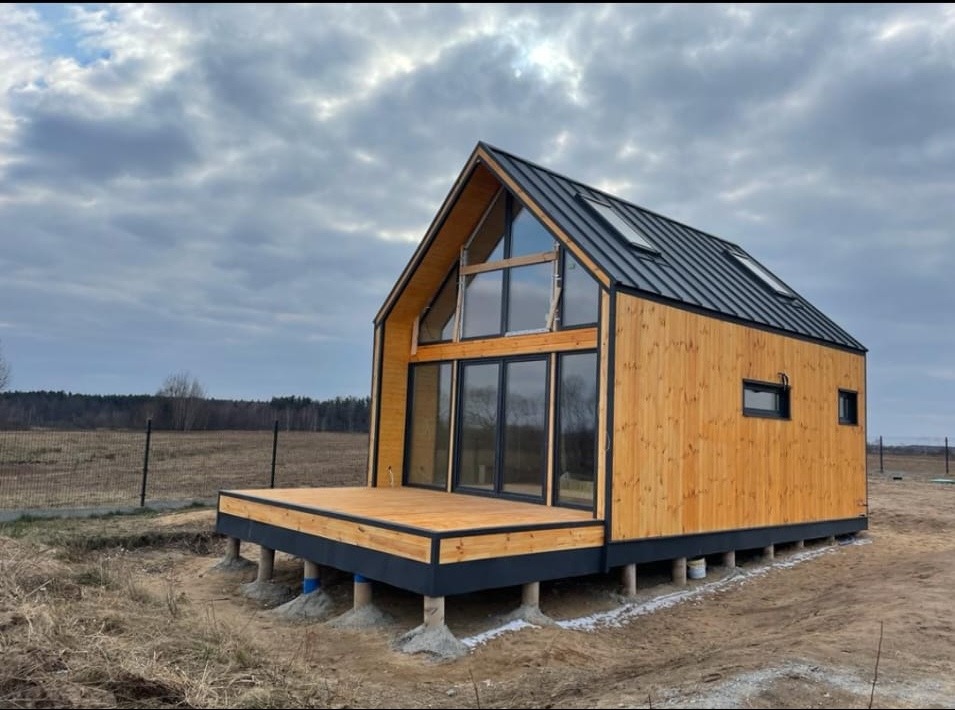 Gotowy, drewniany dom modułowy z dużymi przeszkleniami, posadowiony na betonowych słupach, z tarasem z desek, na tle pochmurnego nieba.