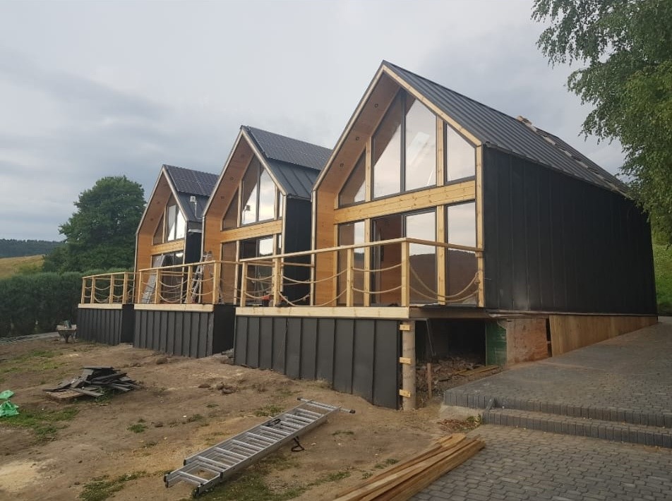 Trzy domy szkieletowe w trakcie budowy, z widocznymi elementami konstrukcyjnymi, drewnianymi balustradami z linami i czarną blachą na elewacji, na nierównym terenie z porozrzucanymi materiałami...