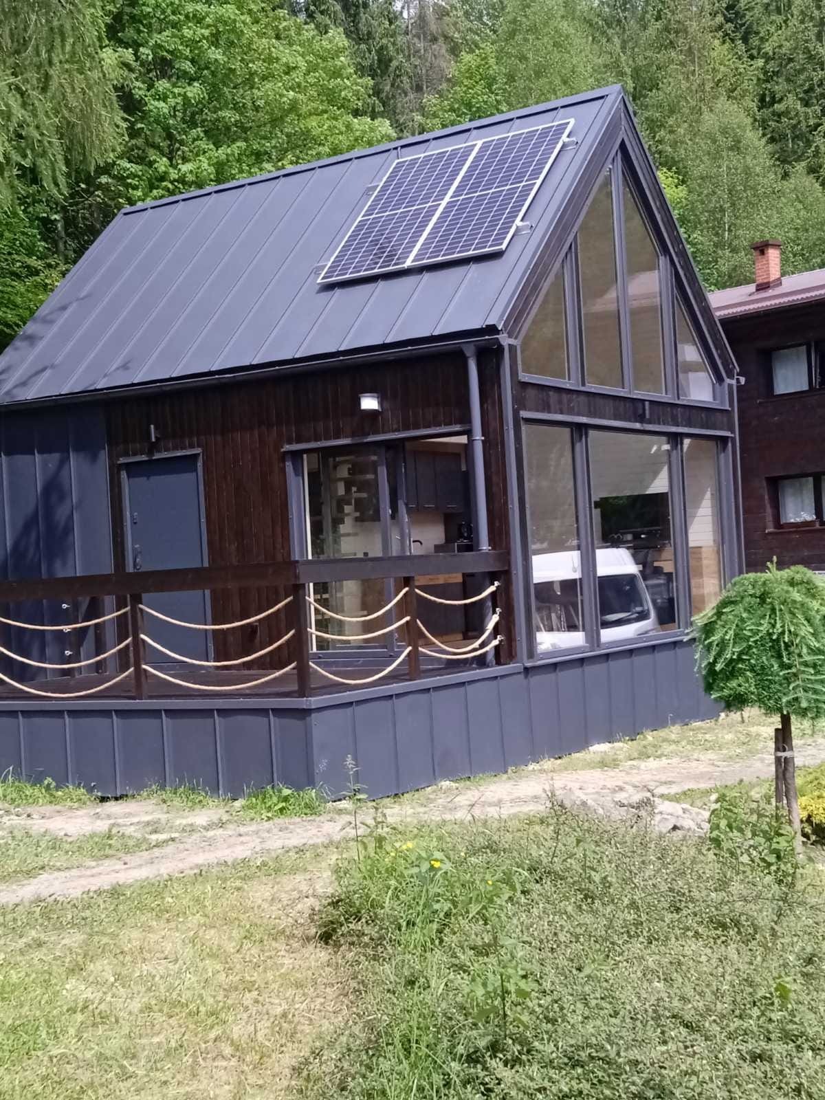 Nowoczesny, ciemny dom z panelami słonecznymi na dachu, z dużymi oknami i drewnianym tarasem z linową balustradą, otoczony zielenią.