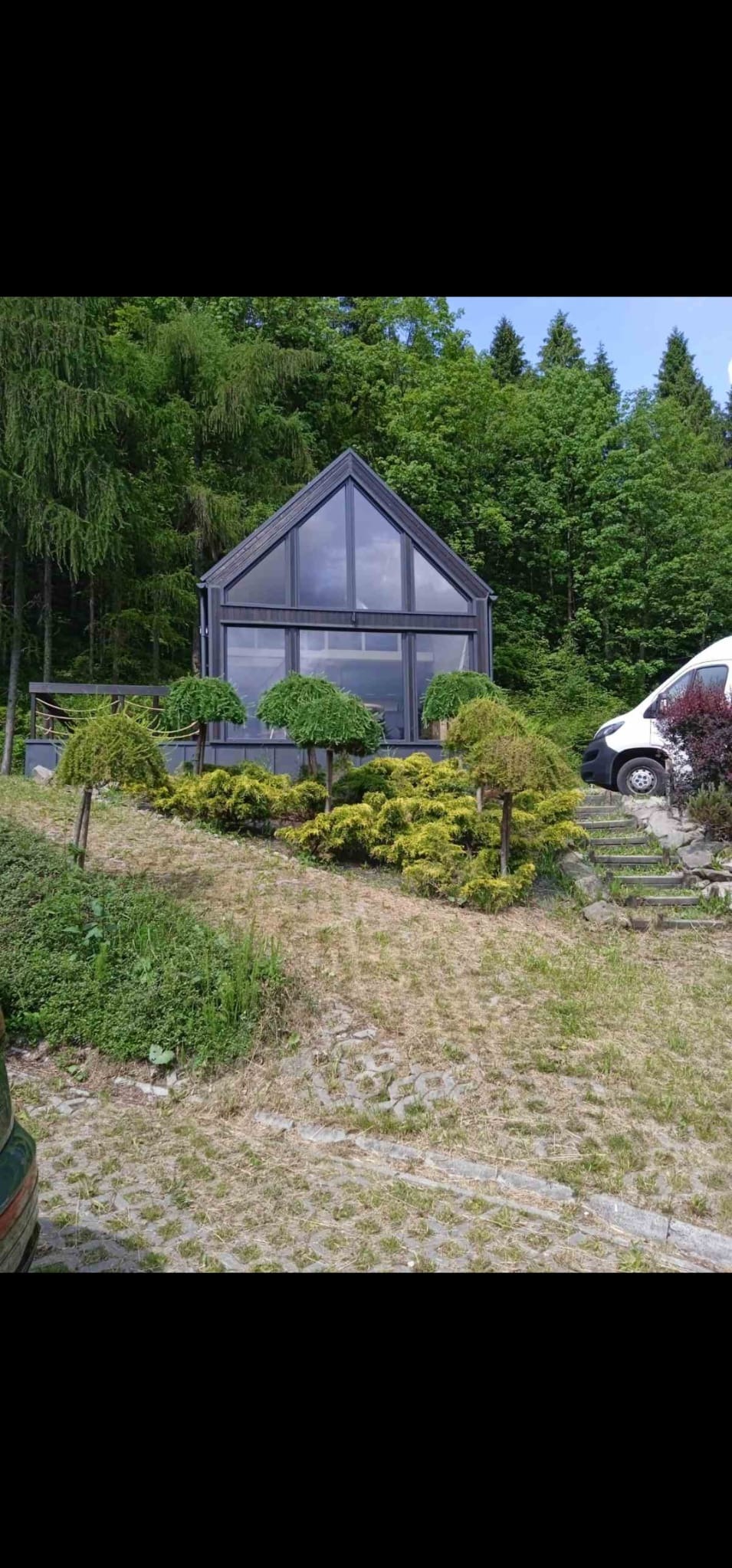 Nowoczesny, przeszklony budynek w stylu stodoły, otoczony zielenią i ozdobnymi krzewami, z częściowo widocznym samochodem dostawczym w tle.