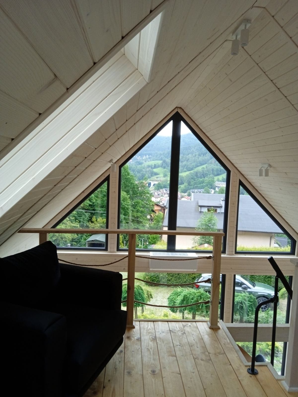 Wnętrze pokoju na poddaszu z drewnianą boazerią, trójkątnym oknem z widokiem na zalesione wzgórza i czarnym fotelem w rogu.