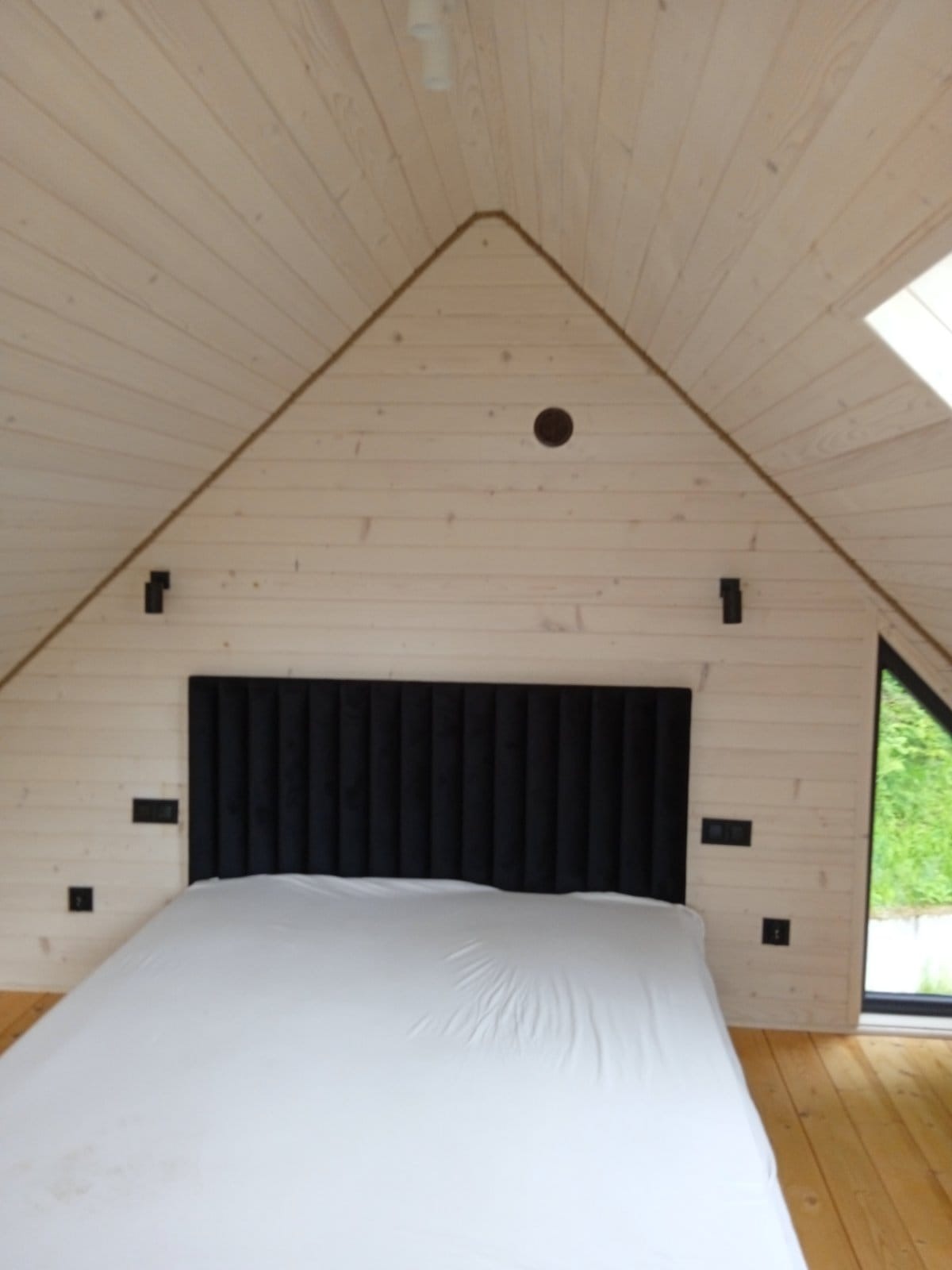 Wnętrze sypialni w domu z drewnianych paneli, z czarnym zagłówkiem i białym prześcieradłem na łóżku, małe okno trójkątne w ścianie szczytowej.
