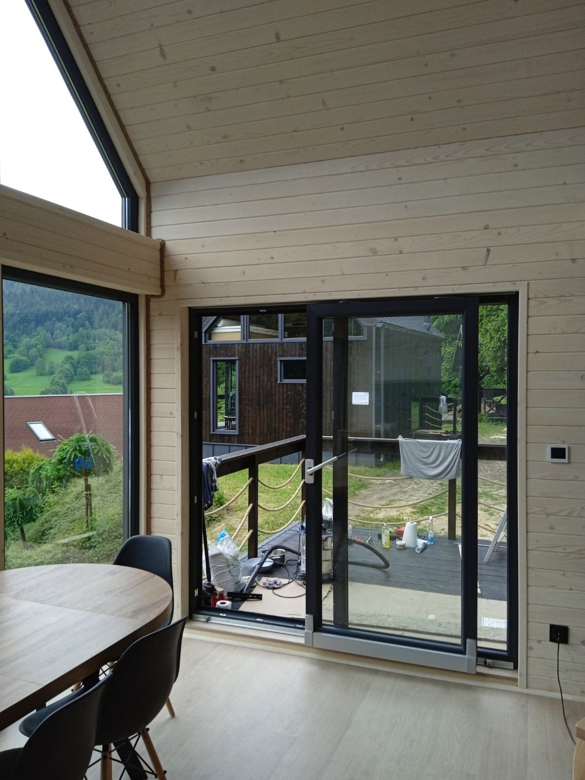 Wnętrze domu z drewnianą boazerią i dużymi przesuwnymi drzwiami balkonowymi, widok na taras i ogród, okrągły stół z krzesłami w rogu pomieszczenia, na podłodze panele.