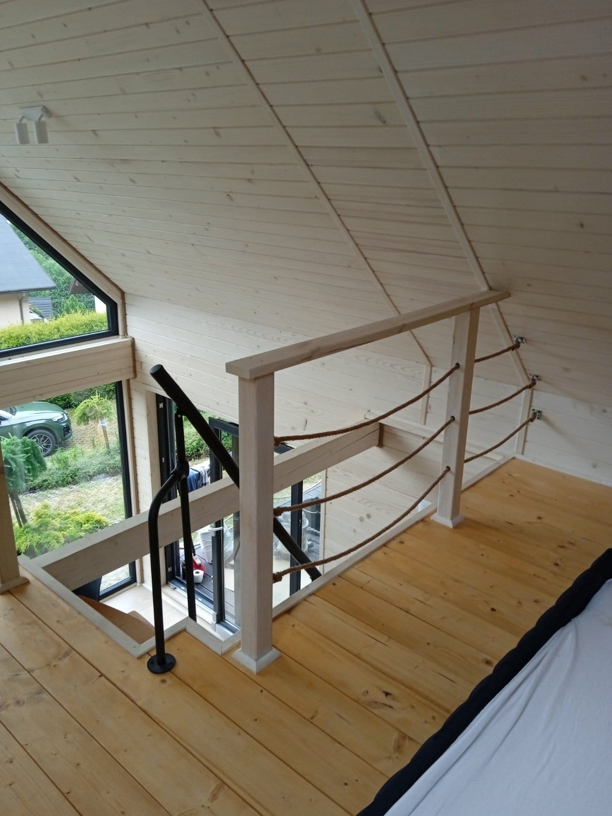 Wnętrze domu szkieletowego z drewnianymi schodami z czarną poręczą i zabezpieczeniem z liny jutowej, widok z antresoli na okno z widokiem na ogród.