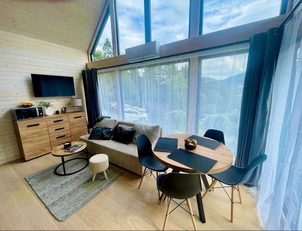 Jasne wnętrze salonu w domu z drewnianą boazerią, duże okno z widokiem na góry, minimalistyczny wystrój z drewnianymi meblami i szarymi akcentami, okrągły stół z owocami.