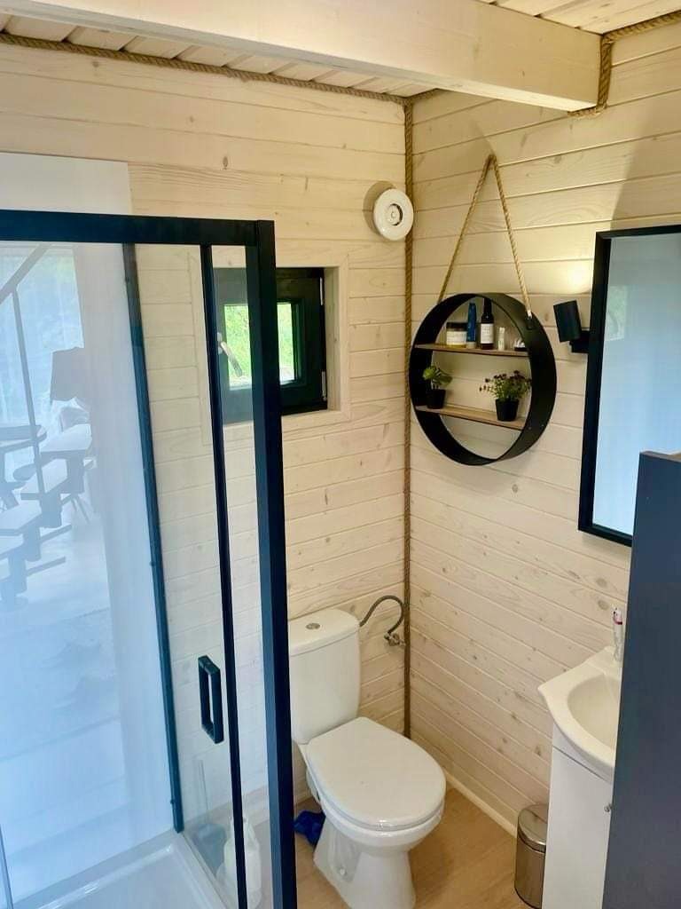 Jasna łazienka z kabiną prysznicową z czarną ramą, toaletą, umywalką i okrągłą półką wiszącą na sznurku, wykończona drewnianą boazerią.