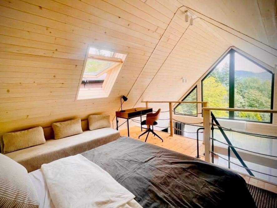 Jasne wnętrze poddasza z drewnianą boazerią, łóżkiem, biurkiem i dużym oknem z widokiem na las.