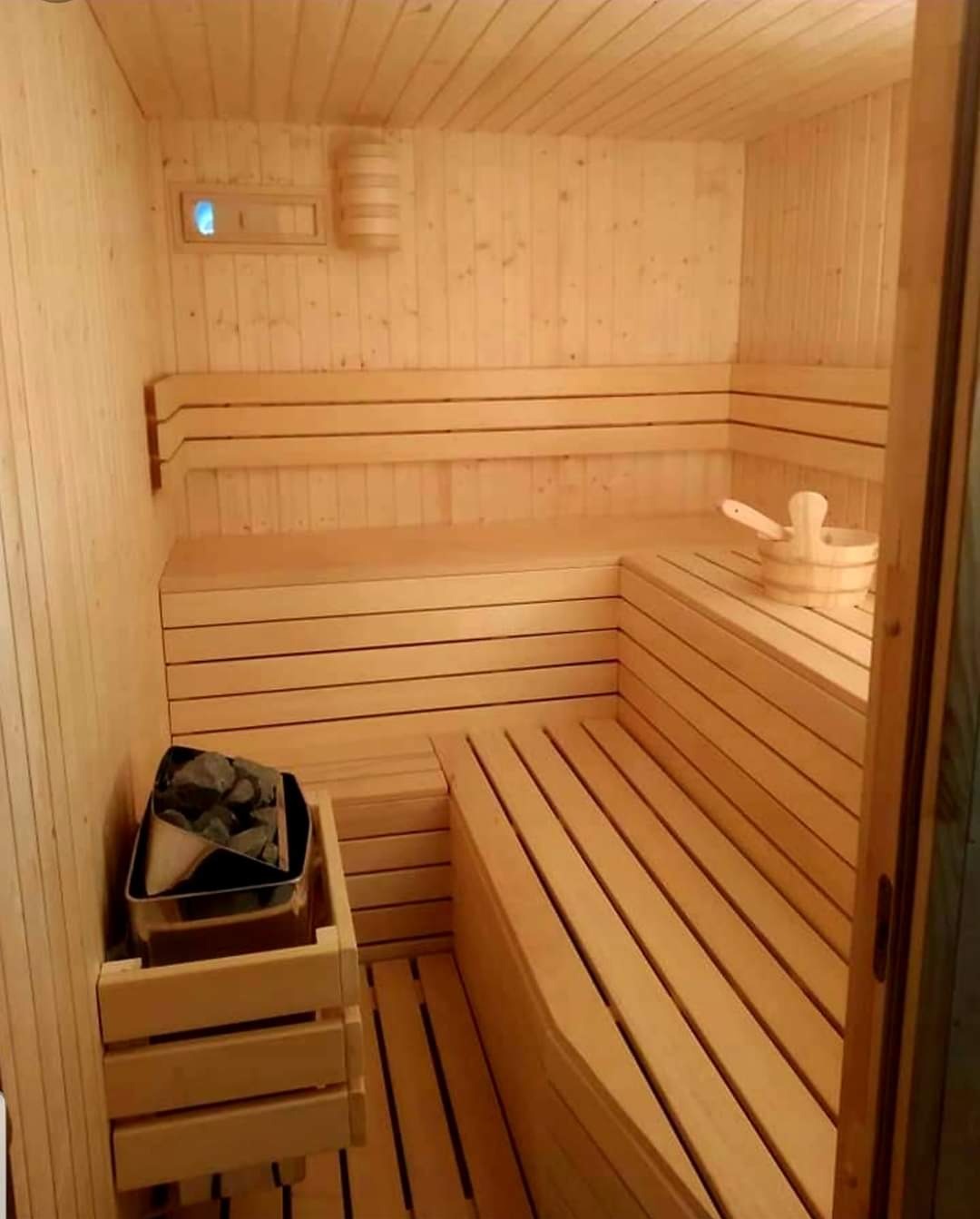 Wnętrze sauny wykończonej jasnym drewnem, z drewnianymi ławkami, pojemnikiem z kamieniami i drewnianym czerpakiem.