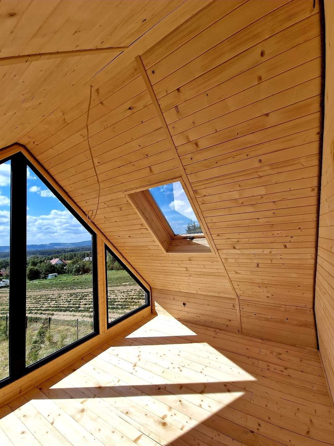 Wnętrze poddasza wykończonego boazerią z widokiem na krajobraz przez duże okno trapezowe i okno dachowe.