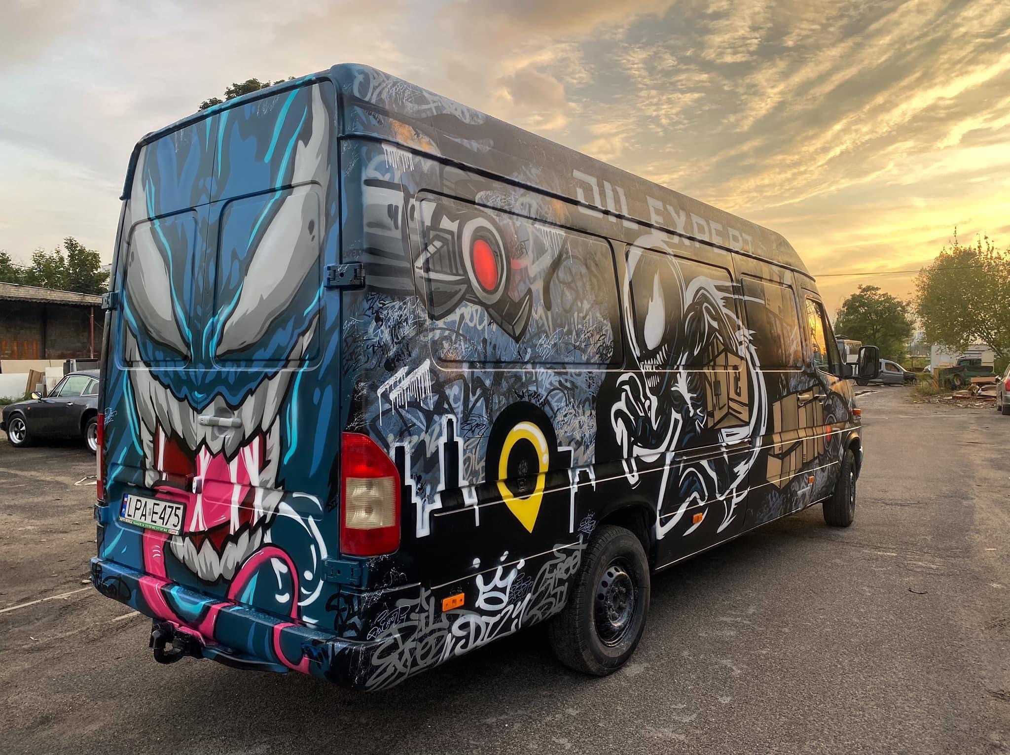 Czarny bus z artystycznym graffiti przedstawiającym potwory i symbole miejskie, zaparkowany na zewnątrz przy zachodzącym słońcu.