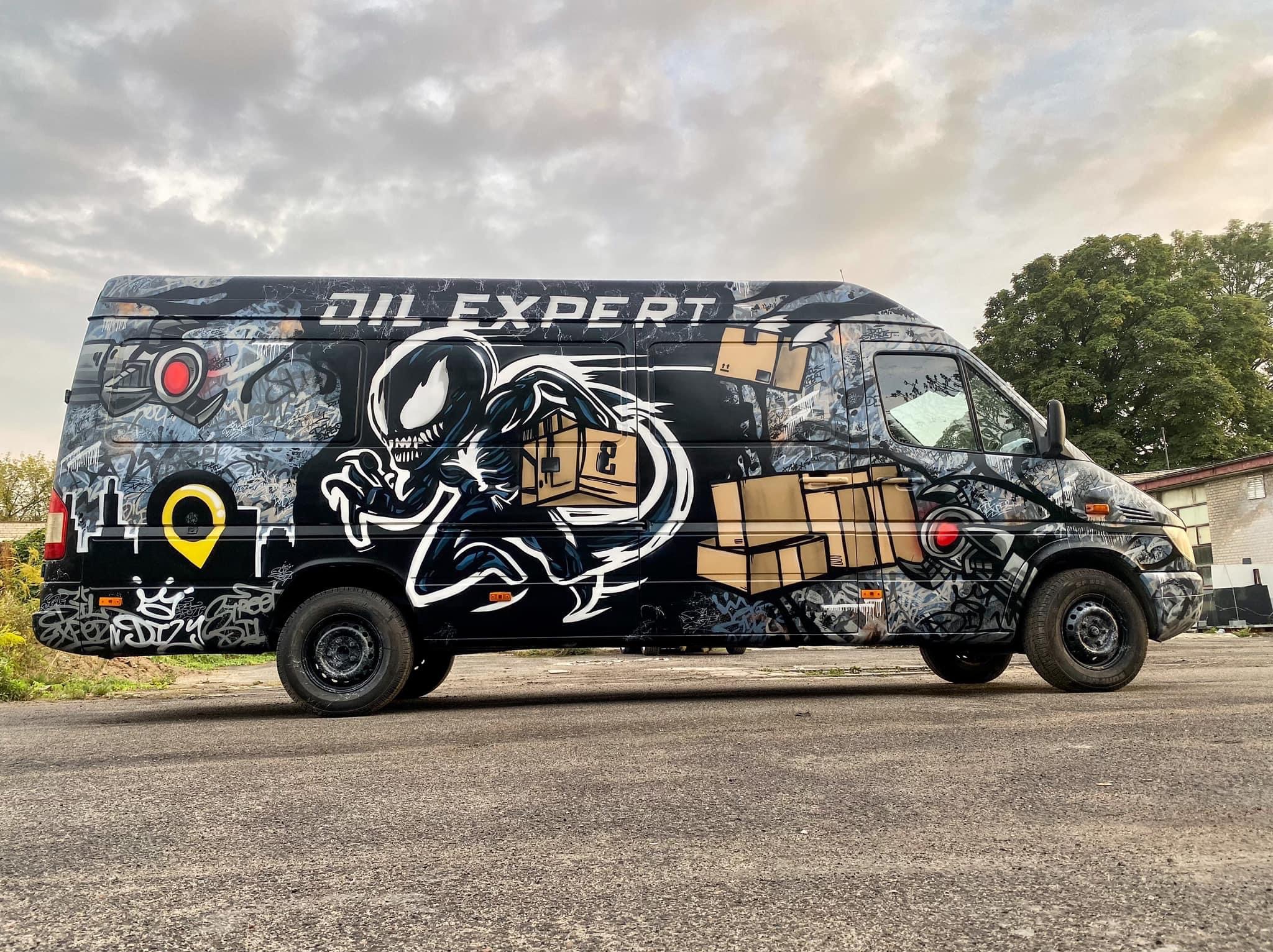 Czarny bus z artystycznym graffiti przedstawiającym postać w stylu Venom niosącą kartony, z napisem 'OIL EXPERT' na boku, zaparkowany na szarym asfalcie pod pochmurnym niebem.