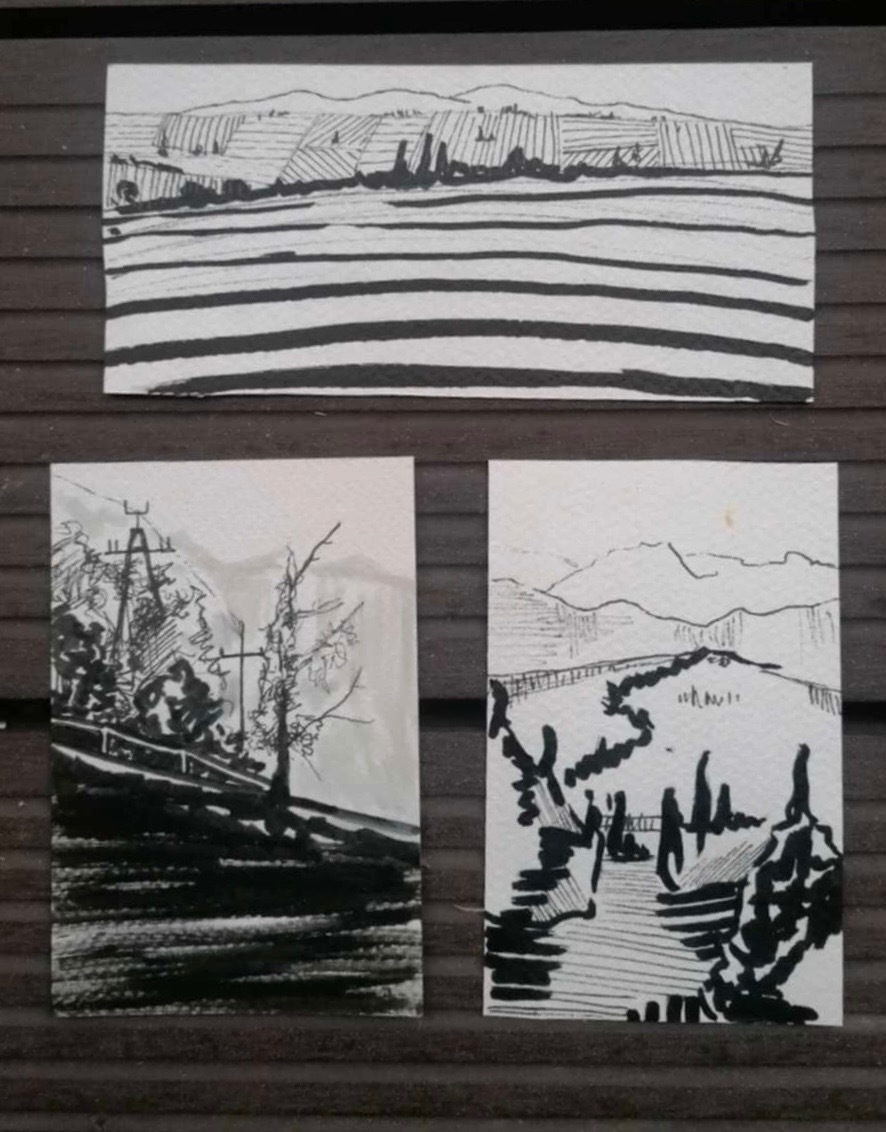 Trzy czarno-białe szkice tuszem przedstawiające krajobrazy: pola z liniami, drzewa przy drodze i ścieżkę w górach, umieszczone na brązowym, drewnianym tle.