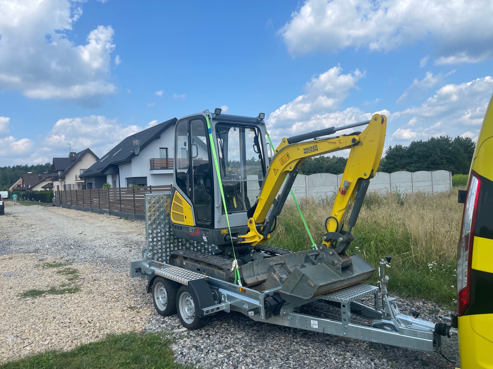 Żółto-szara mini koparka Wacker Neuson ET18, zabezpieczona zielonymi pasami, transportowana na przyczepie dwuosiowej z metalową platformą, na tle domu jednorodzinnego i błękitnego nieba z chmurami.