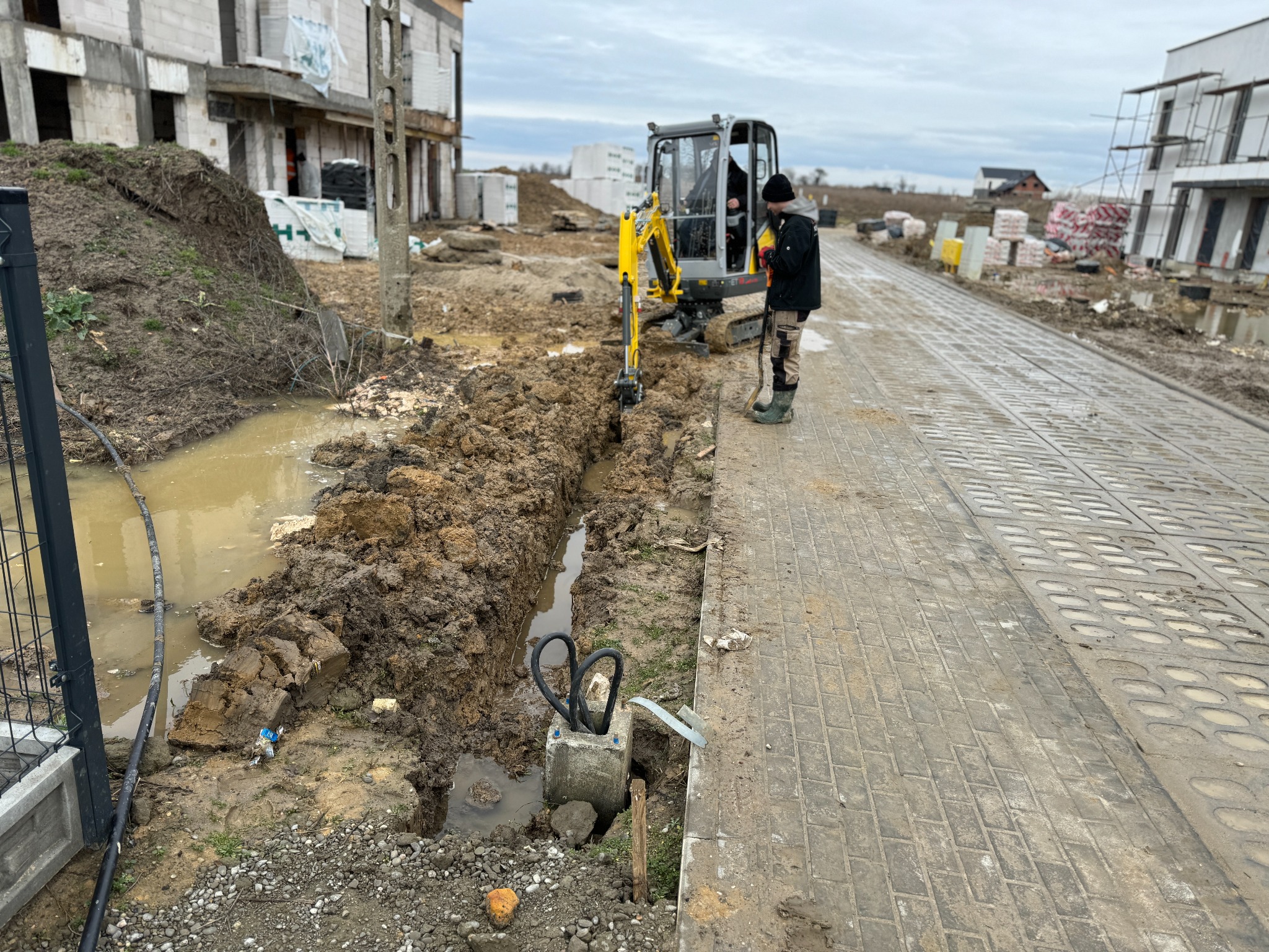 Żółta mini koparka podczas wykonywania wąskiego wykopu w błotnistym gruncie na placu budowy, widoczny operator w kabinie i fragment ułożonej drogi z płyt betonowych.