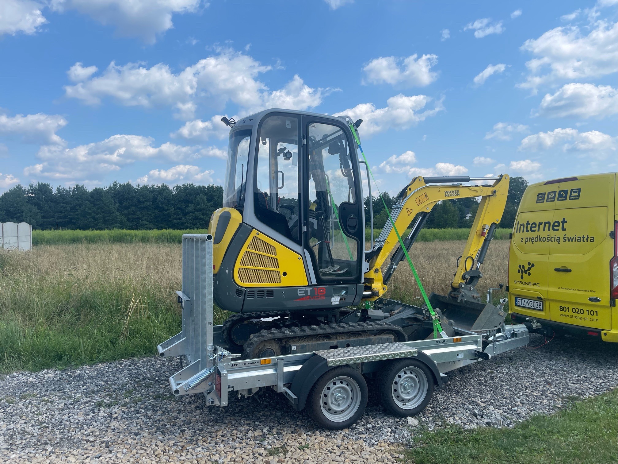 Żółto-szara mini koparka Wacker Neuson ET18, zabezpieczona zielonymi pasami, transportowana na srebrnej przyczepie, obok żółtego samochodu dostawczego z napisem 'Internet z prędkością światła'...