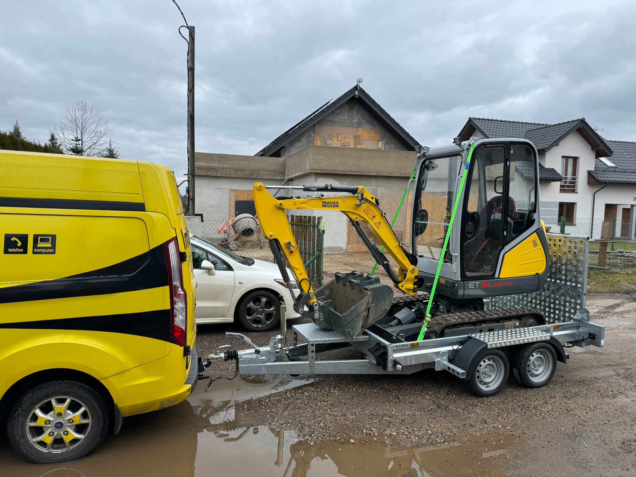Żółto-czarny bus ciągnie na przyczepie mini koparkę Wacker Neuson ET18, teren podmokły, w tle budynek w trakcie budowy i dom mieszkalny.
