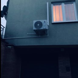 JohnnyKlima JAN GAJEWSKI - Zewnętrzny agregat klimatyzacji GREE zamontowany na szarej ścianie budynku, okno z odbiciem zachodu słońca, antena satelitarna i kamera przemysłowa w tle. Ujęcie z dołu.