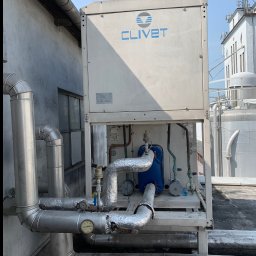 JohnnyKlima JAN GAJEWSKI - Biały agregat wody lodowej Clivet na dachu budynku, z widocznymi rurami instalacji chłodniczej, manometrami i elementami hydraulicznymi. W tle budynki i anteny.