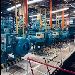 JohnnyKlima JAN GAJEWSKI - Rząd niebieskich, przemysłowych agregatów chłodniczych HS1-HS3 z pomarańczowymi i żółtymi rurami w hali technologicznej, widok zza barierki.