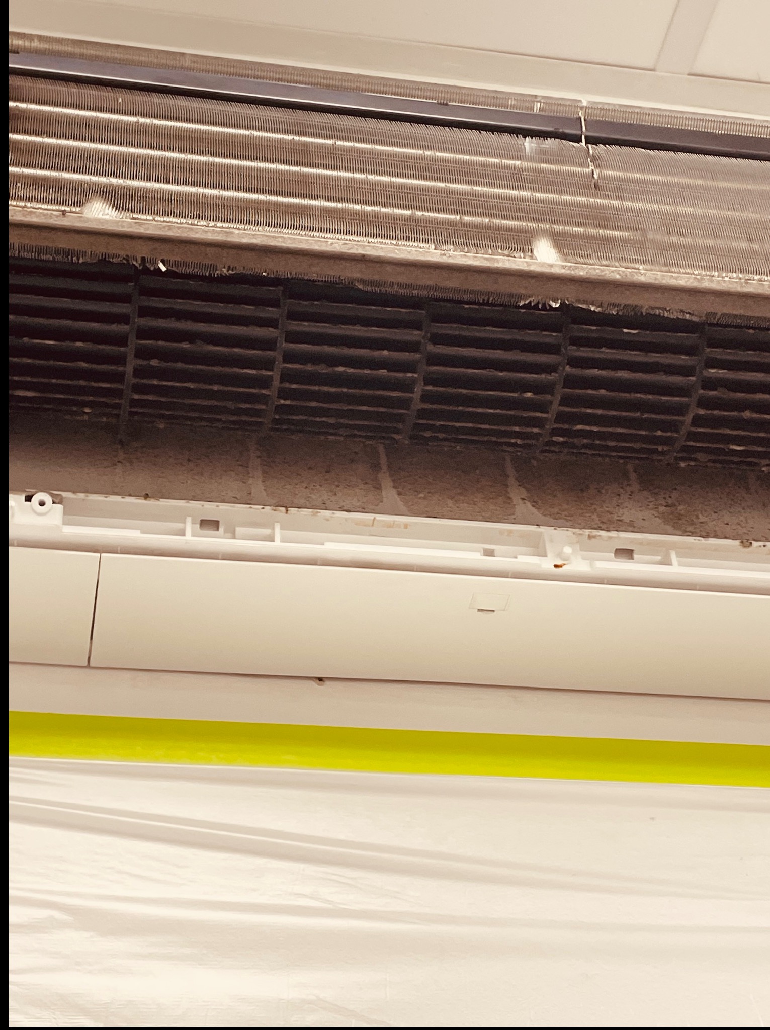 Zanieczyszczony, otwarty klimatyzator z widocznymi filtrami i wentylatorem pokrytymi kurzem, przygotowany do czyszczenia, z zabezpieczonym podłożem folią malarską.