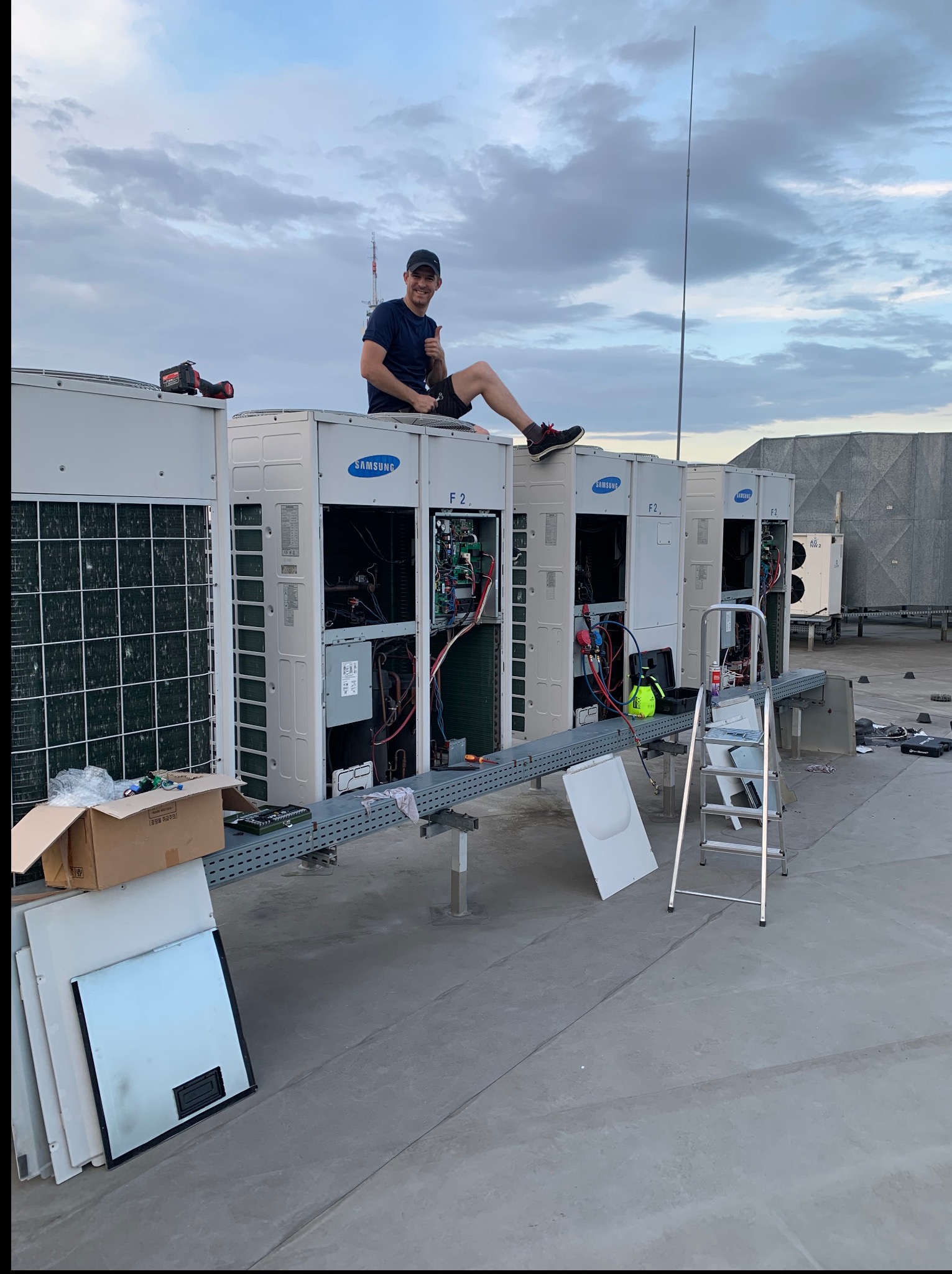 Instalacja klimatyzacji Samsung na dachu budynku. Mężczyzna siedzi na urządzeniu, dając znak kciukiem w górę. Widoczne narzędzia i drabina.