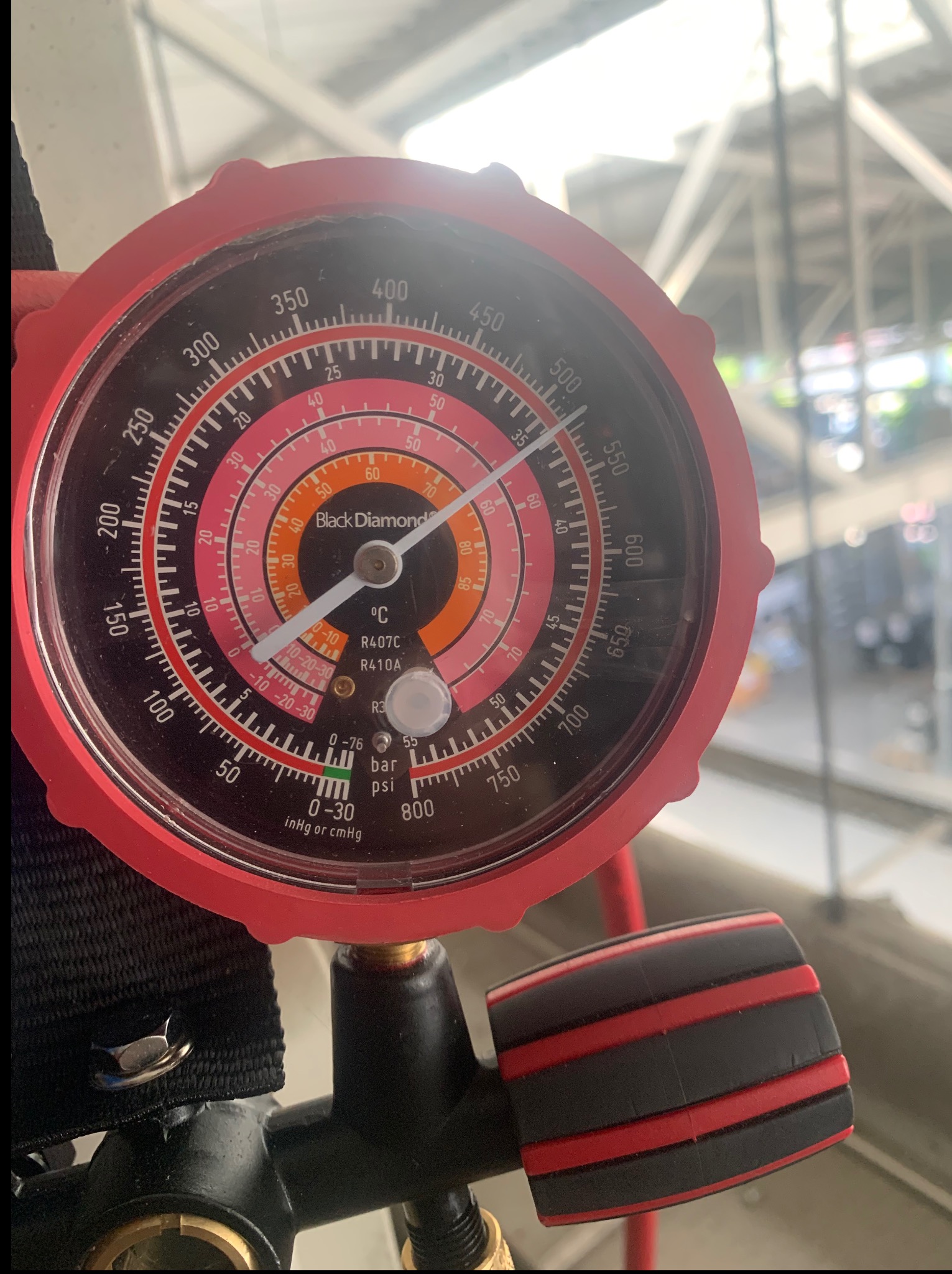 Manometr Black Diamond z czerwoną obudową, wskazujący ciśnienie czynnika chłodniczego R407C/R410A, na tle konstrukcji stalowej. Widoczne złącze i fragment węża.