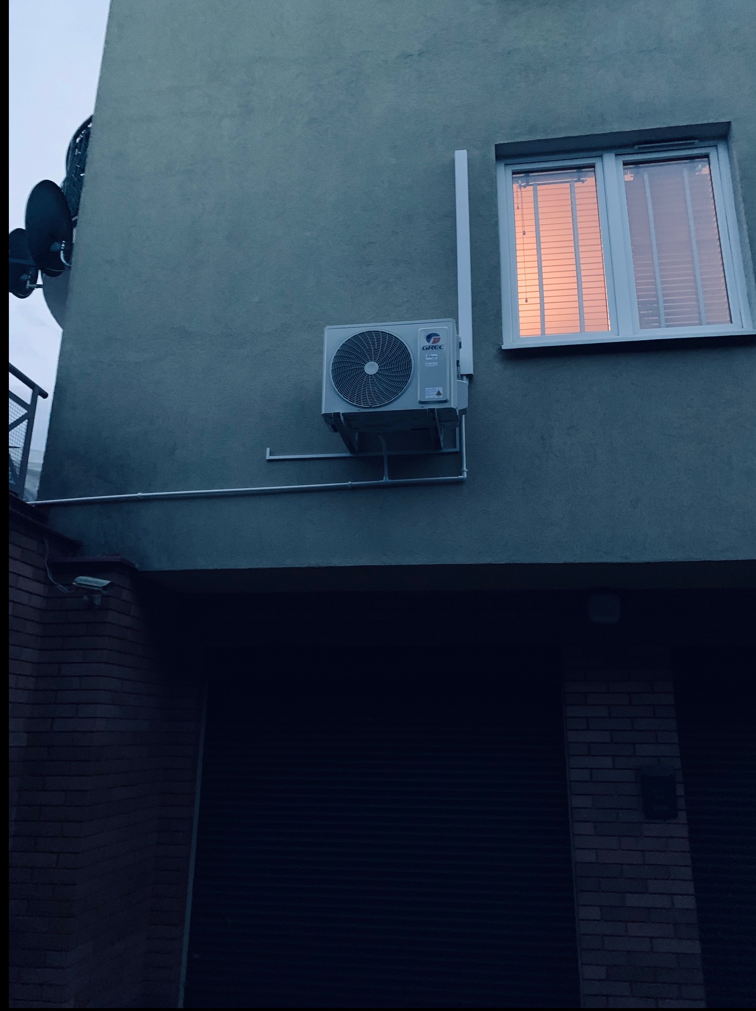 Zewnętrzny agregat klimatyzacji GREE zamontowany na szarej ścianie budynku, okno z odbiciem zachodu słońca, antena satelitarna i kamera przemysłowa w tle. Ujęcie z dołu.