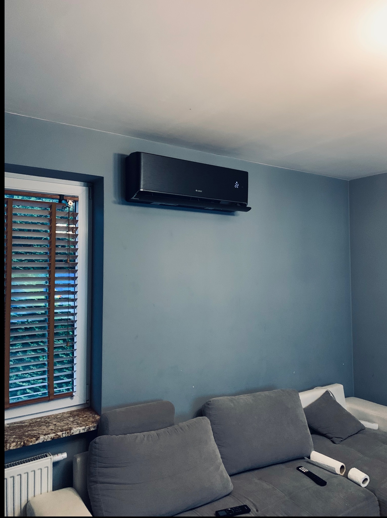 Czarny klimatyzator Gree zamontowany na szarej ścianie w pokoju, obok okna z drewnianymi żaluzjami i szarą sofą. Widoczny wyświetlacz temperatury na urządzeniu.