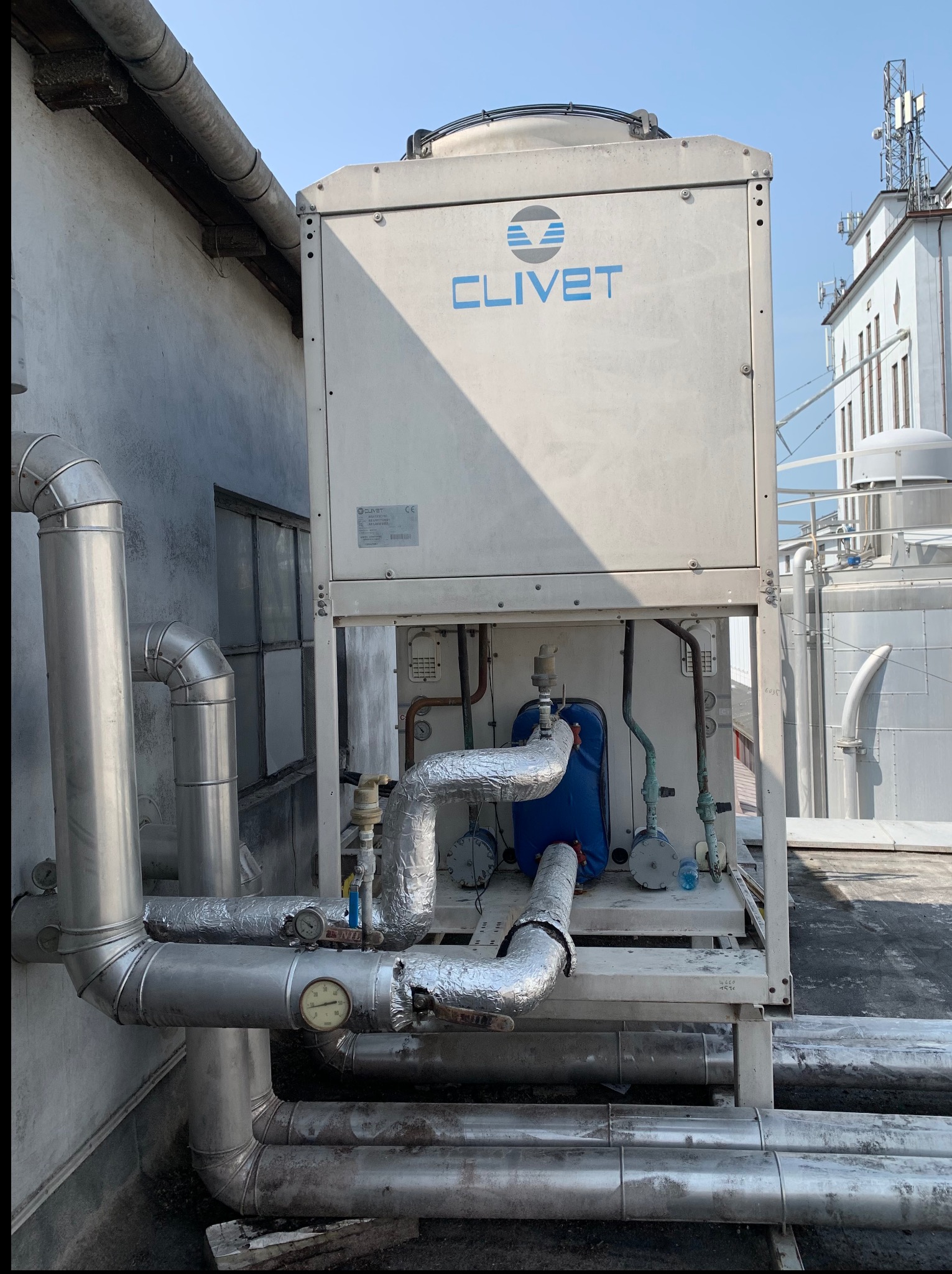 Biały agregat wody lodowej Clivet na dachu budynku, z widocznymi rurami instalacji chłodniczej, manometrami i elementami hydraulicznymi. W tle budynki i anteny.