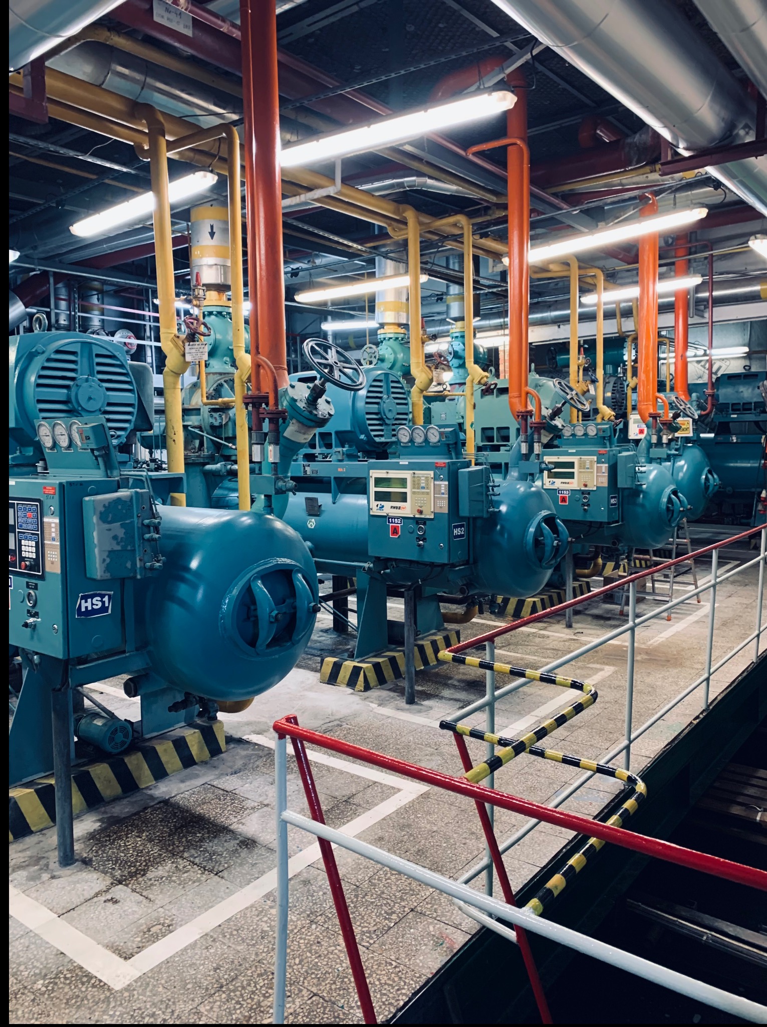 Rząd niebieskich, przemysłowych agregatów chłodniczych HS1-HS3 z pomarańczowymi i żółtymi rurami w hali technologicznej, widok zza barierki.