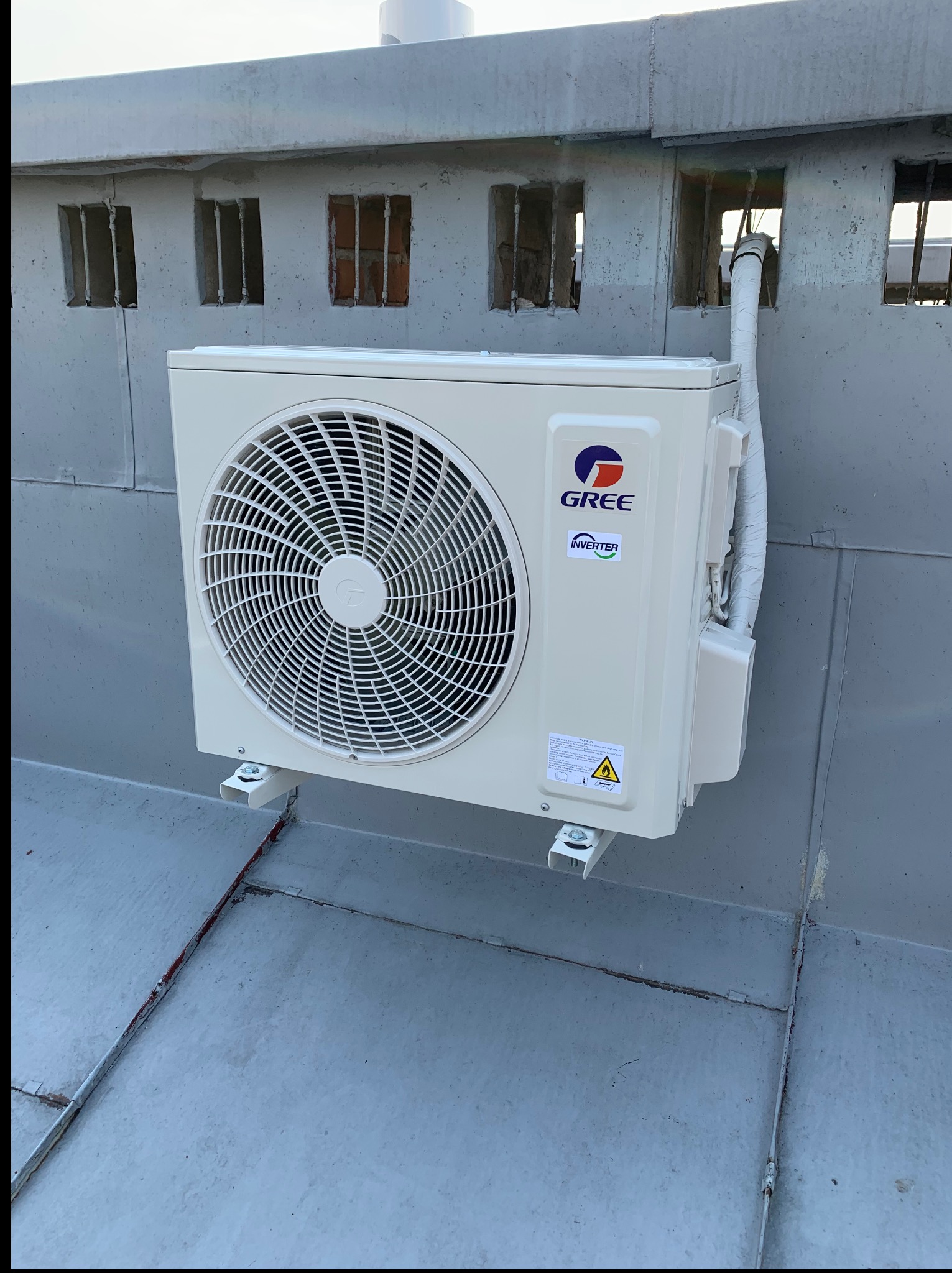 Biała jednostka zewnętrzna klimatyzacji Gree, zamontowana na szarym dachu budynku z małymi, zakratowanymi oknami w tle. Widoczne rury i przewody.