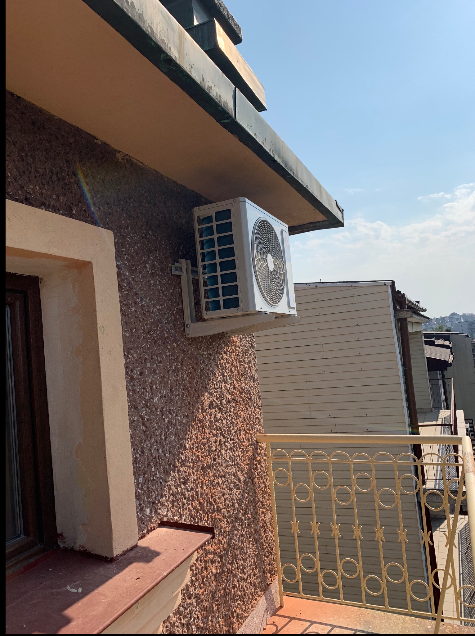 Biała klimatyzacja na balkonie budynku z teksturą, widok z dołu na błękitne niebo. Metalowa balustrada balkonu i sąsiednie budynki w tle.