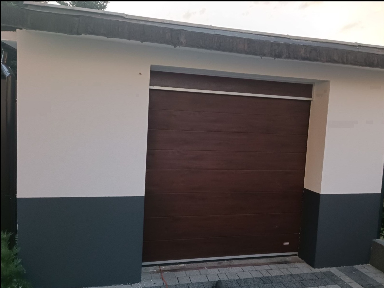 Świeżo pomalowany garaż z brązową bramą segmentową i dwukolorową elewacją: górna część biała, dolna część szara.