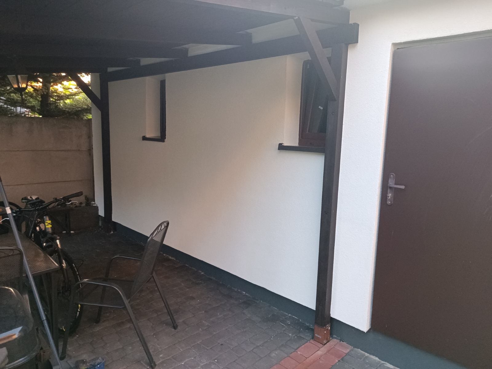 Świeżo otynkowana ściana domu pod zadaszeniem, z widocznymi oknami i ciemnymi elementami konstrukcyjnymi, na pierwszym planie rower i krzesło.