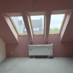 Poddasze z trzema oknami dachowymi, ściany wyłożone płytami karton-gips w kolorze r&oacute;żowym, grzejnik zabezpieczony folią, surowa podłoga.
