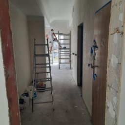 KrzyśBud - Remont korytarza: mężczyzna szpachluje ścianę na rusztowaniu, widoczne drabiny, wiaderka z farbą i zabezpieczone drzwi. Korytarz w trakcie prac wykończeniowych.