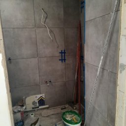 KrzyśBud - Szare płytki ścienne w trakcie układania z widocznymi krzyżykami dystansowymi, puszkami elektrycznymi i narzędziami. Fragment instalacji elektrycznej i wiadro z farbą.