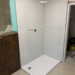 KrzyśBud - Biały brodzik w narożniku łazienki z częściowo wykończonymi ścianami. Widoczne przygotowania pod montaż armatury i fragment nieotynkowanej ściany.