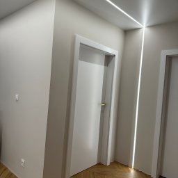 KrzyśBud - Nowoczesny korytarz z oświetleniem LED wzdłuż sufitu i ścian, białe drzwi, parkiet w jodełkę. Minimalistyczny design, jasne wnętrze, świeżo po remoncie.